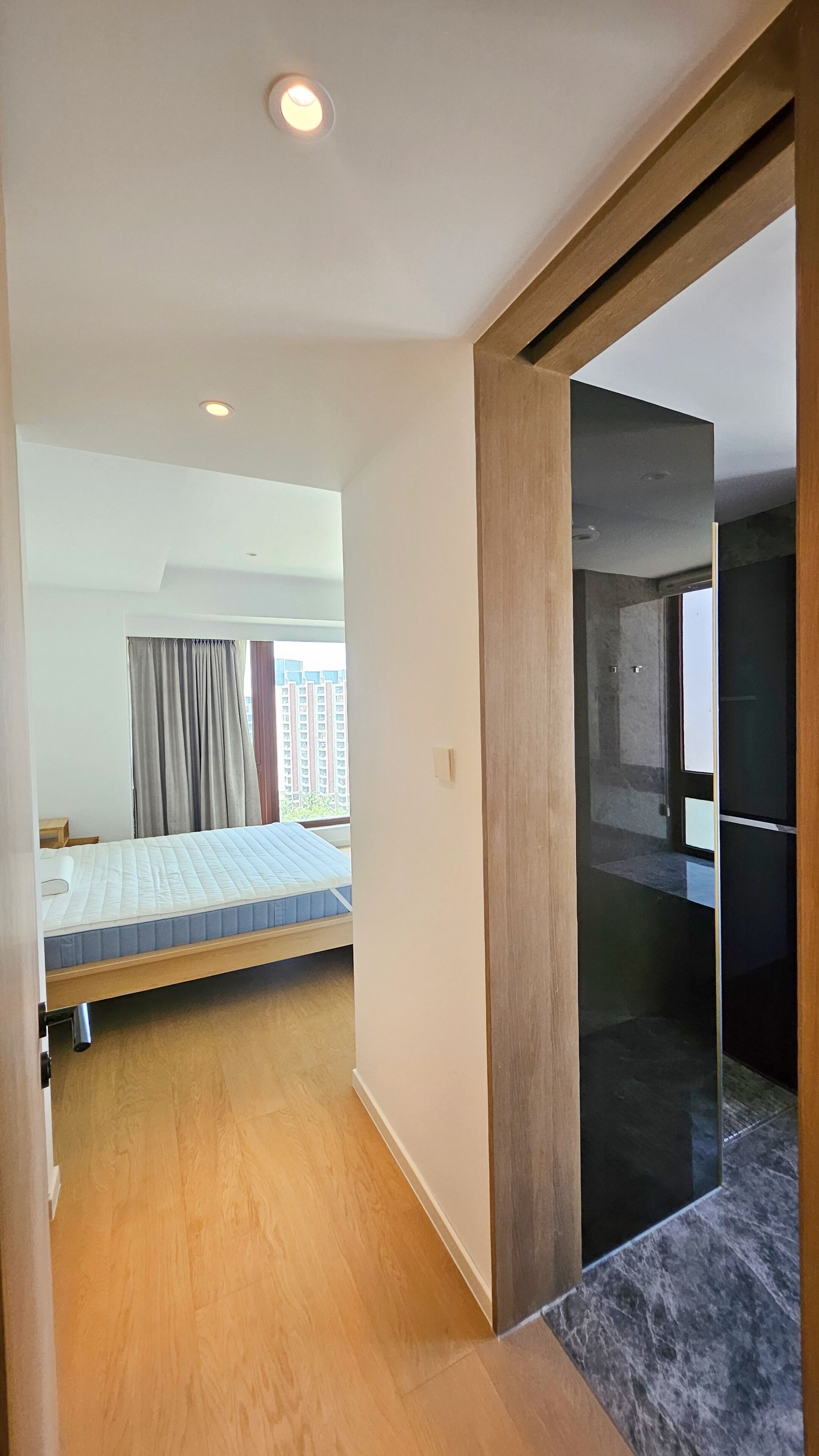 A hotel room with a bed, a wooden floor, a bathroom with a mirror, and a view of buildings outside.