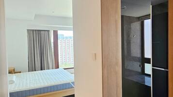 A hotel room with a bed, a wooden floor, a bathroom with a mirror, and a view of buildings outside.