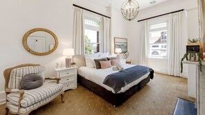 A bedroom with a bed, a chair, a bedside table, a lamp, a mirror, and a window with curtains. - QPO in Historic Queenscliff (Queenscliffe)