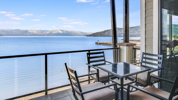 Un balcón con una mesa y sillas de vidrio, con vista a un lago y montañas al fondo.