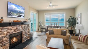 Una sala de estar con chimenea de piedra, un televisor grande, un ventilador de techo y vistas al mar a través de amplios ventanales.