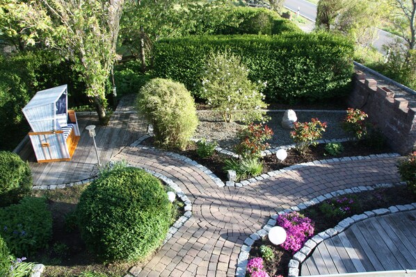 A landscaped garden with a winding brick pathway, neatly trimmed hedges, and a small structure with a sloped roof.