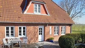 Et murstenshus med et tag af teglsten, en veranda med et bord og stole, samt en velholdt have.