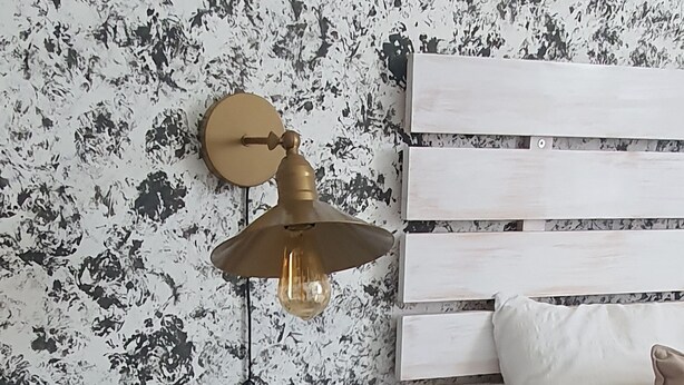 Ein Schlafzimmer mit einem weißen Bett, einem Holzkopfteil, einem kleinen weißen Nachttisch mit einer Topfpflanze und einer Wandlampe.