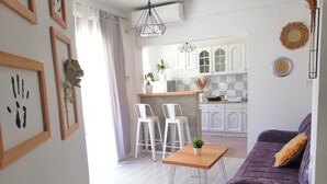 Ein modernes Wohnzimmer mit einem violetten Sofa, weißen Stühlen und einem Holztisch. Im Hintergrund ist eine Küche mit weißen Schränken und einer hängenden Leuchte zu sehen.