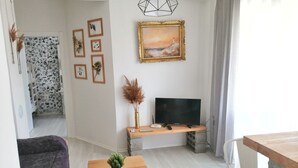 Ein Wohnzimmer mit einer Couch, einem Couchtisch mit Pflanze, einem Fernseher auf einem Schrank und gerahmten Bildern an der Wand.