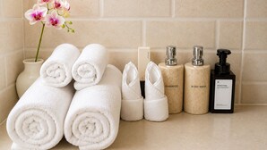 Un tocador de baño con toallas blancas enrolladas ordenadamente, un florero con flores y artículos de aseo, incluyendo champú, gel de baño y jabón de manos.