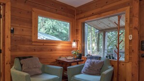 A cozy cabin interior with two upholstered chairs, a small table with books and a plant, and a window with a view of trees. - Cozy cabin in the Woods, Hot tub, secluded, walk to ZigZag River ! (Rhododendron)