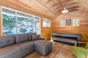 Interior acogedor de una cabaña con un sofá seccional, un sillón verde, techo de madera y ventanas amplias con vistas a un paisaje nevado.