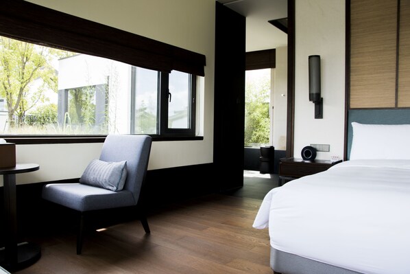 A modern hotel room with a large bed, a seating area with a chair, a small table, and a window offering a view of the outdoors.