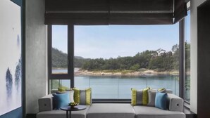 Una habitación moderna con un ventanal grande con vistas a un lago y colinas. Hay un sofá con cojines y una pequeña mesa redonda con una planta.