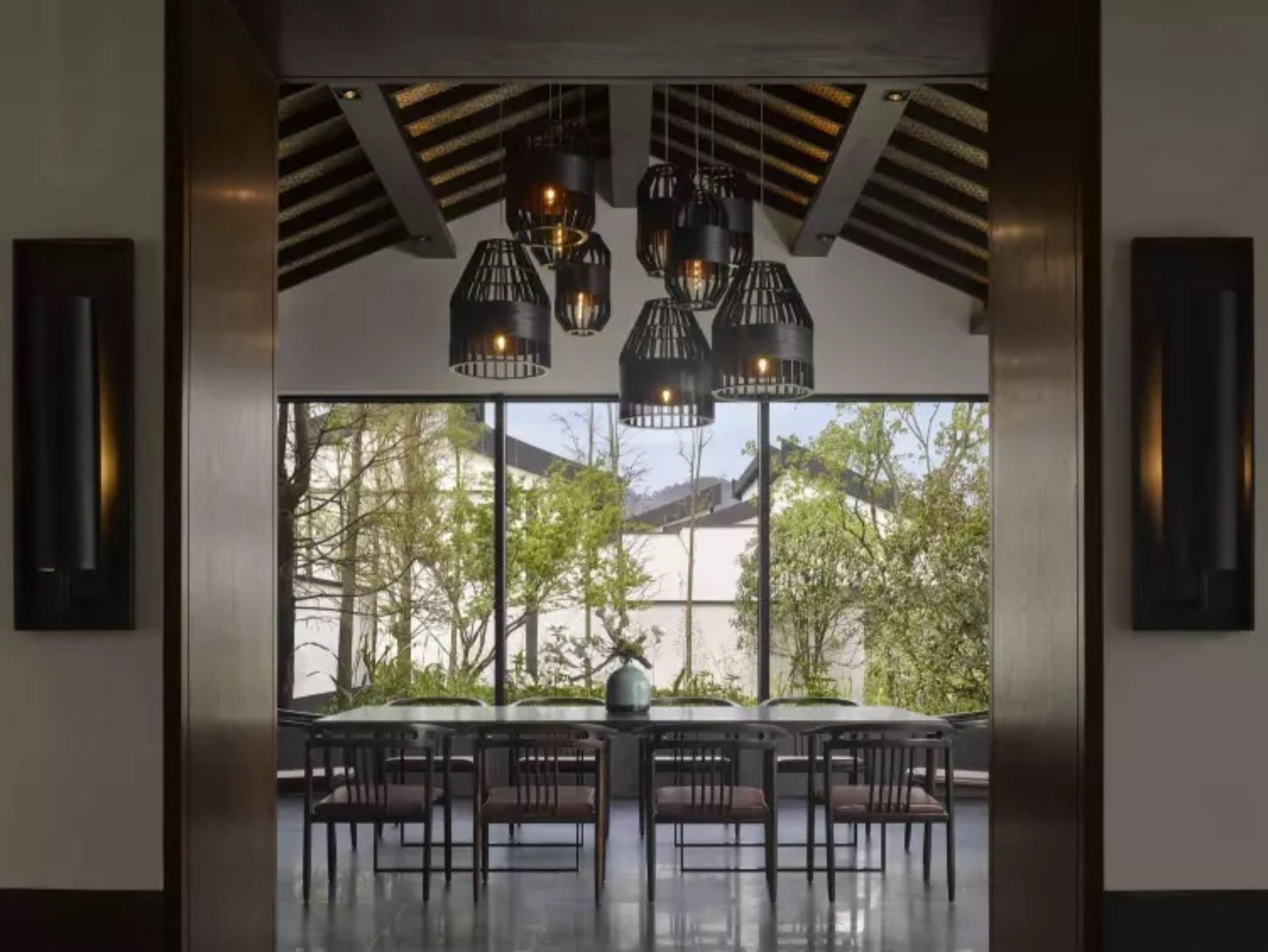 Un comedor moderno con una mesa grande y sillas, ventanas amplias con vistas a árboles y un edificio, y lámparas colgantes.