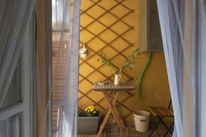 Un petit balcon avec un mur en treillis jaune, une table en bois sur laquelle est posée une carafe blanche, une plante en pot et un seau.