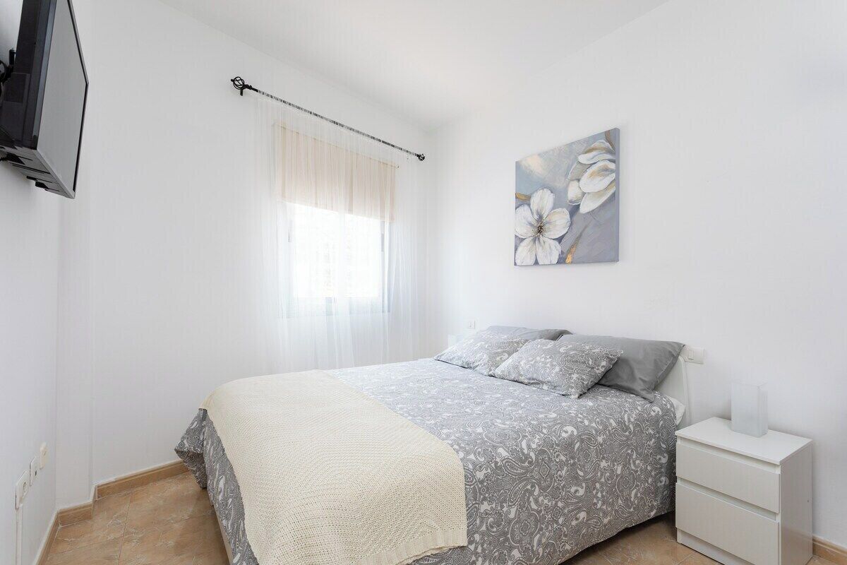 A bedroom with a bed, a bedside table, a TV mounted on the wall, and a window with curtains.