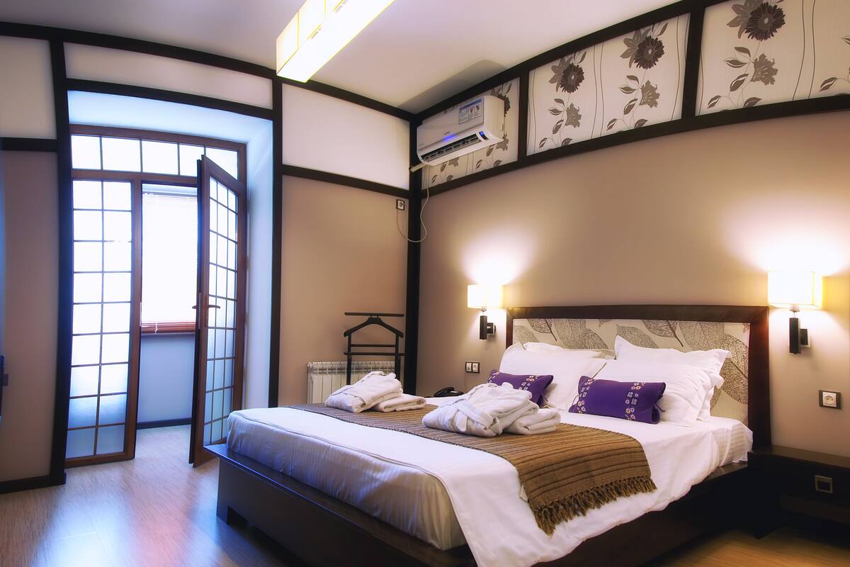 Habitación de hotel con una cama grande, dos lámparas de noche, un radiador y vistas a través de una puerta corredera.