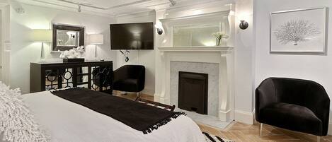 A bedroom with a large bed, a fireplace, a TV, and a desk with a lamp.