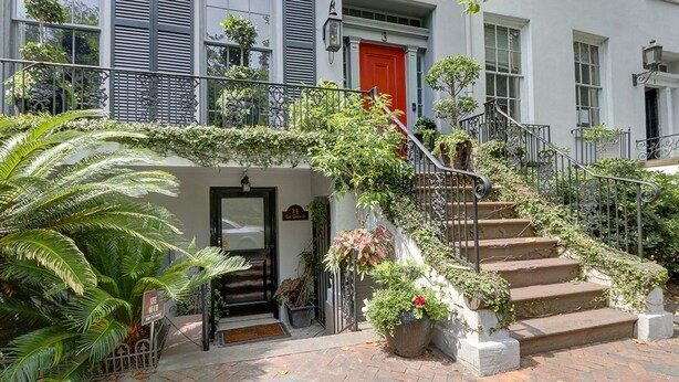 A building with a red door, a staircase with a wrought iron railing, and a small garden area with potted plants. - Heated Pool Access | 5*Clean | Flex Cancelation (Savannah)