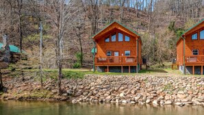 Two wooden cabins with balconies, a stone retaining wall, and a calm lake. - Pigeon Forge River Front Cabin- Fishing Year Round-firepit with seating and Hottub (Pigeon Forge)