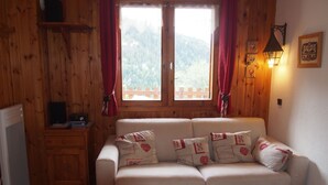 Ein gemütliches Zimmer mit Holzvertäfelung, einem weißen Sofa mit gemusterten Kissen, einem kleinen Tisch und einem Fenster mit Vorhängen.