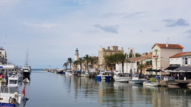 A marina with boats docked along a waterfront, buildings with balconies, and a prominent tower structure. - Very bright villa standing 5 minutes walk from the beach  (Le Grau-du-Roi)