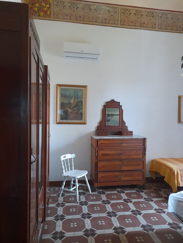 A room with a patterned floor, a wooden dresser, a chair, a bed with a yellow bedspread, and a wall-mounted air conditioner.