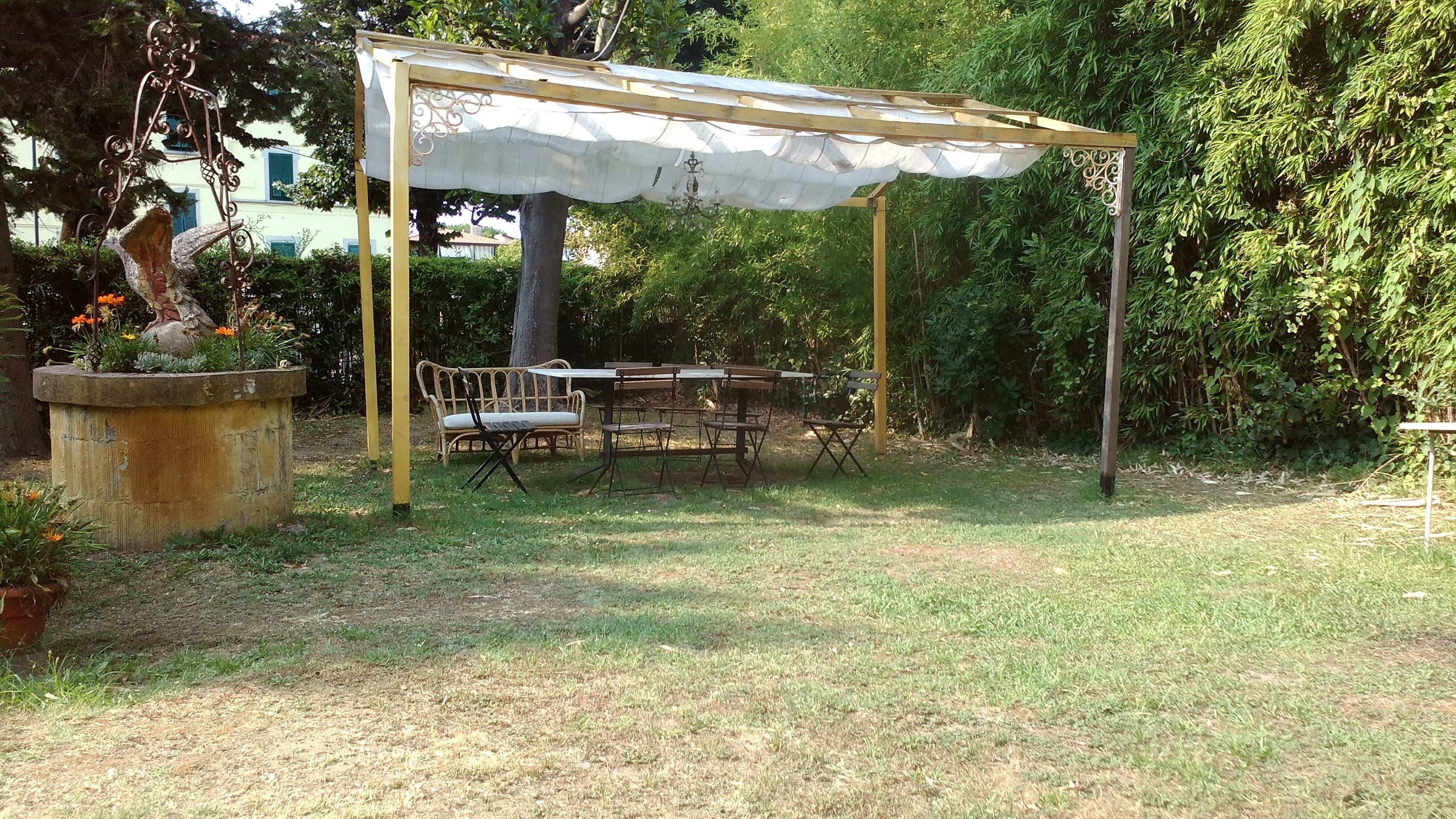 A covered outdoor seating area with a table and chairs, surrounded by greenery and a concrete planter with flowers.