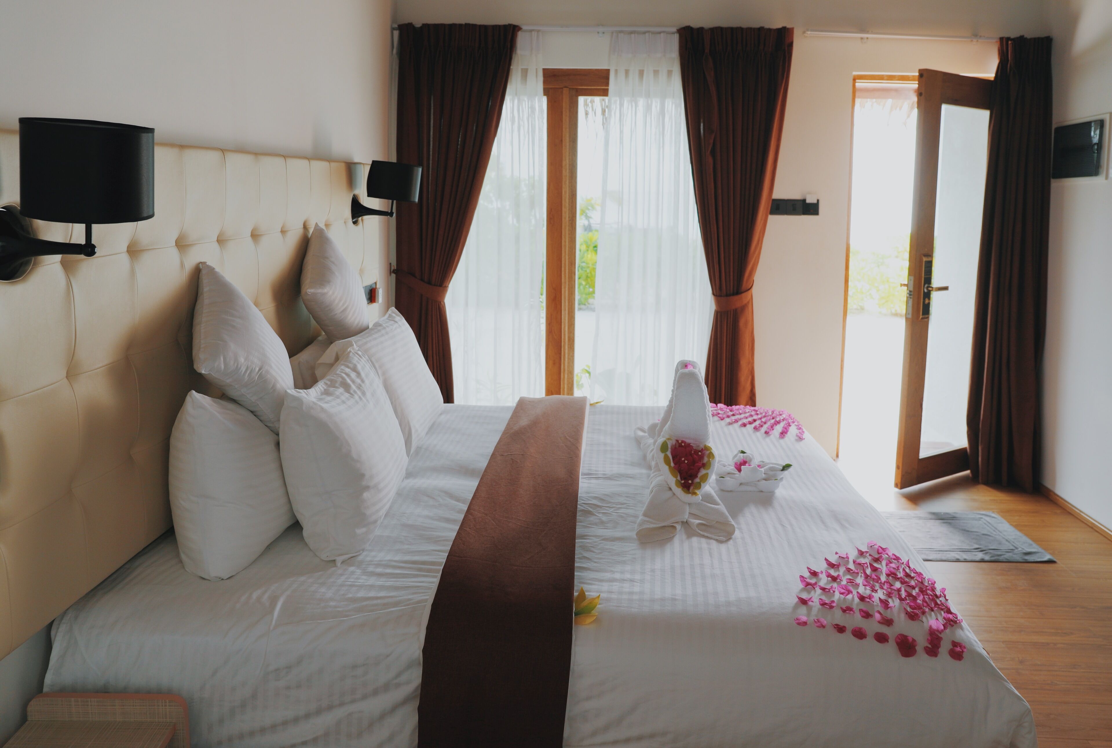 A neatly made bed with white linens, a brown bedspread, and a bouquet of flowers on the pillow.