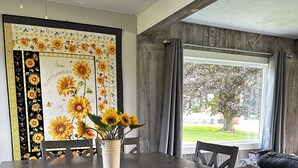 Un comedor con una mesa y sillas de madera, un jarrón con girasoles y un amplio ventanal con cortinas.
