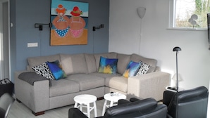 Una sala de estar con un sofá seccional gris, una mesa de centro blanca y un sillón negro. Hay un cuadro en la pared y una lámpara de pie.