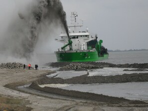 A green ship with a white superstructure, docked at a rocky shore with a group of people standing nearby.