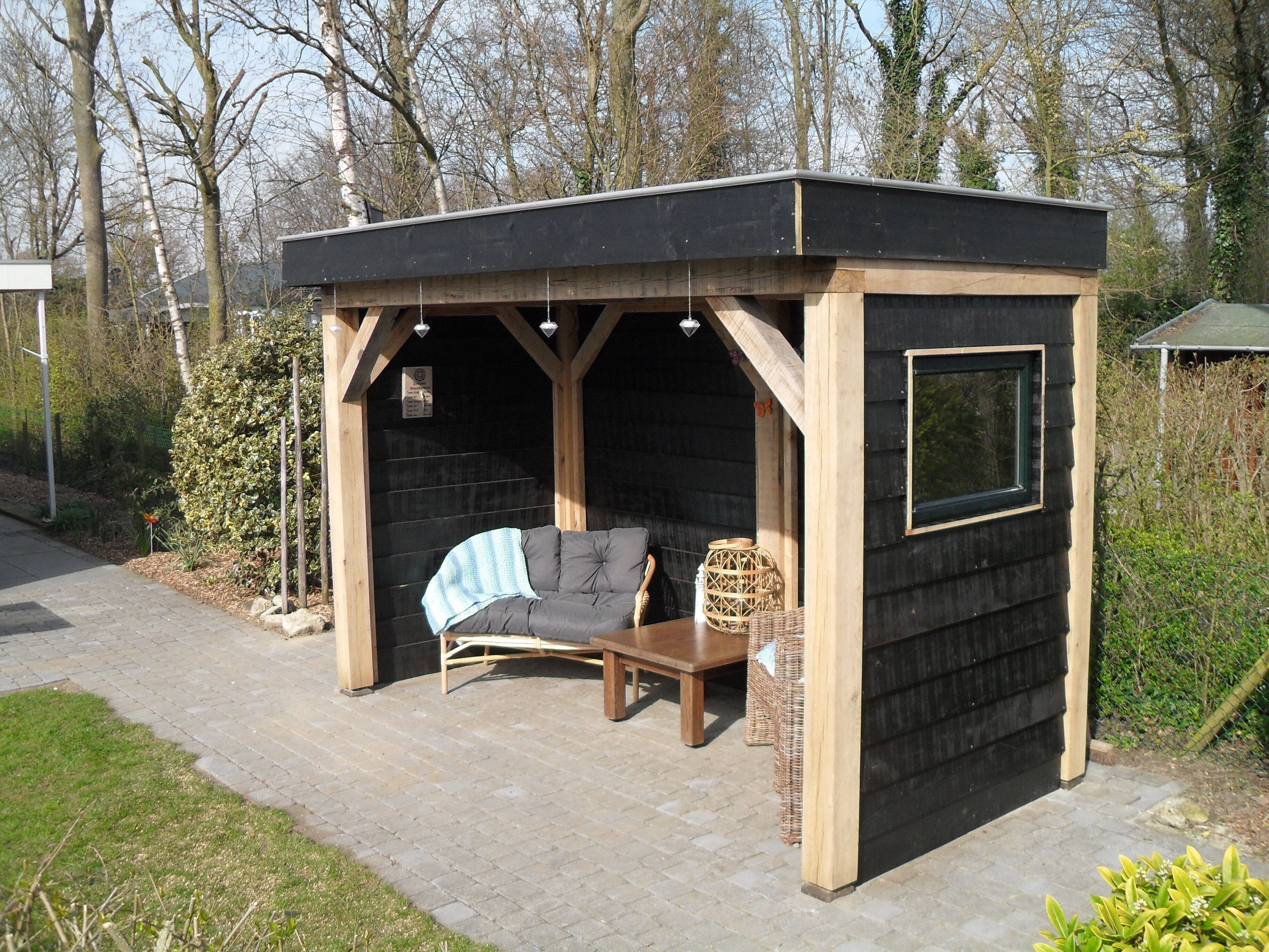 Een houten prieel met een zithoek met kussens, een tafeltje en een raam.