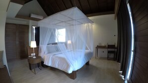 Une chambre à coucher avec un lit recouvert d’une moustiquaire, une table de chevet avec une lampe, une armoire en bois et une fenêtre avec des rideaux.