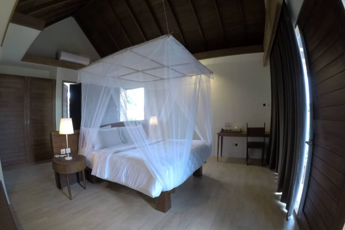 A bedroom with a bed under a mosquito net, a bedside table with a lamp, a wooden wardrobe, and a window with curtains.