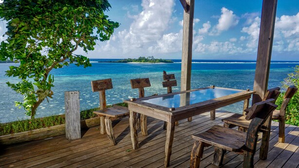 Une terrasse en bois avec une table et des chaises à plateau de verre, donnant sur une plage tropicale aux eaux d’un bleu limpide et sur une île lointaine.