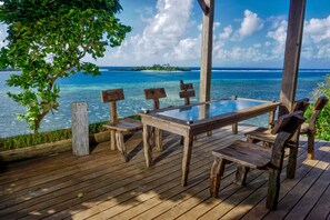 Een houten terras met een tafel en stoelen met glazen blad, met uitzicht op een tropisch strand met helderblauw water en een eiland in de verte.