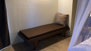 Une petite chambre avec un lit simple, un banc en bois, un coussin et un luminaire fixé au mur.