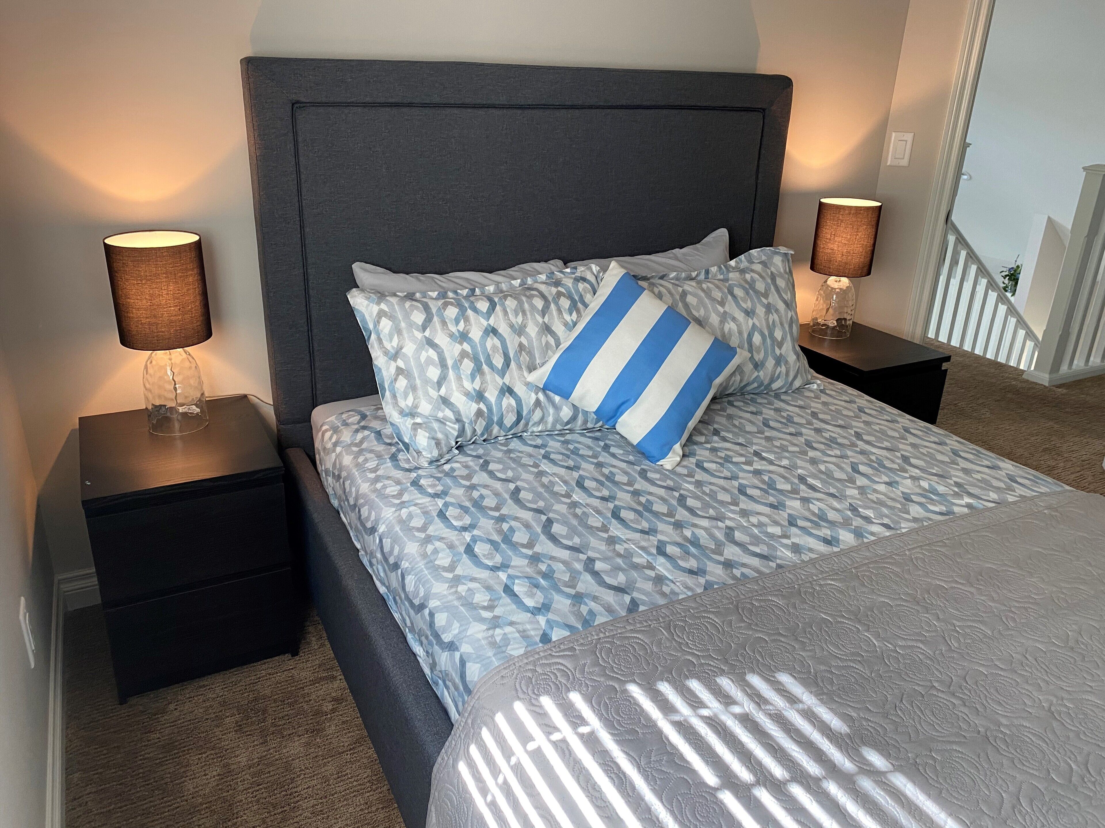 Een slaapkamer met een grijs gestoffeerd hoofdeinde, een bed met een dessin en twee bedlampjes.