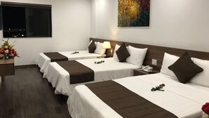 A hotel room with three beds, each with white linens and brown headboards. There is a painting on the wall, a window with a view, and a small table with a vase of flowers.