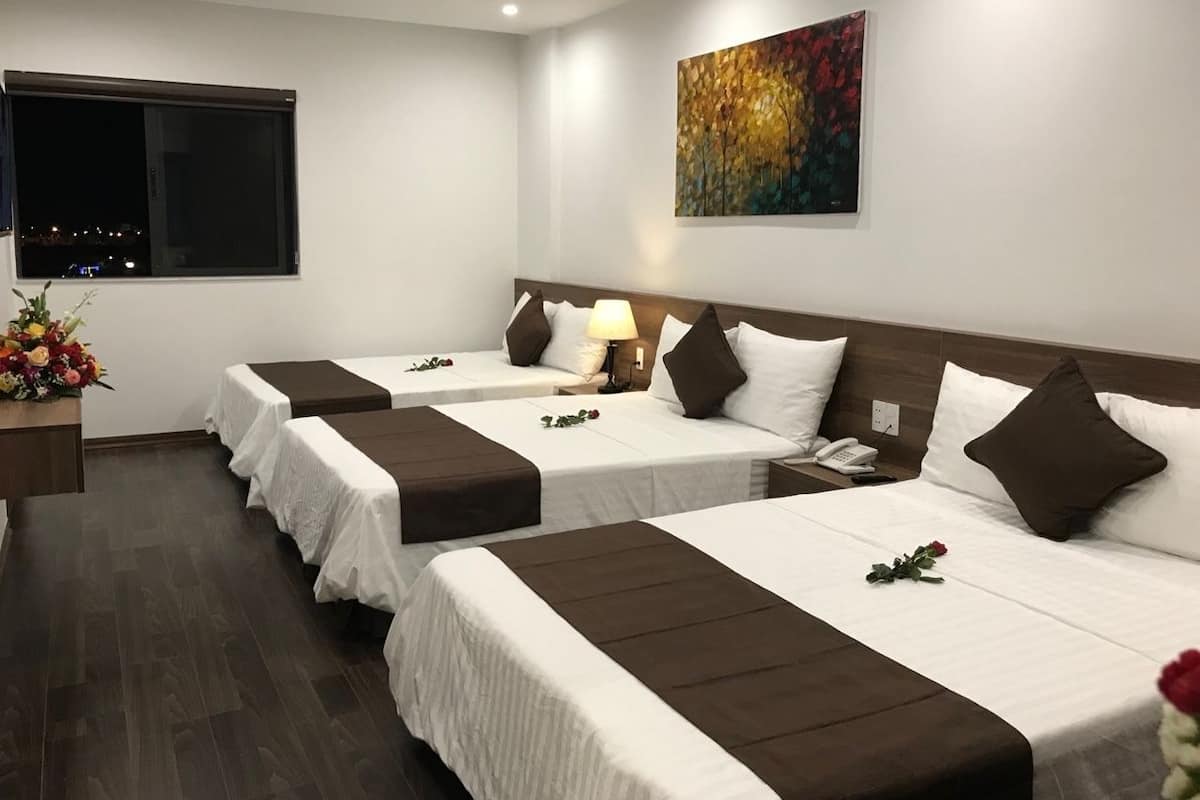 Quarto de hotel com três camas, cada uma com roupa de cama branca e cabeceiras castanhas. Há uma pintura na parede, uma janela com vista e uma mesa pequena com um vaso de flores.