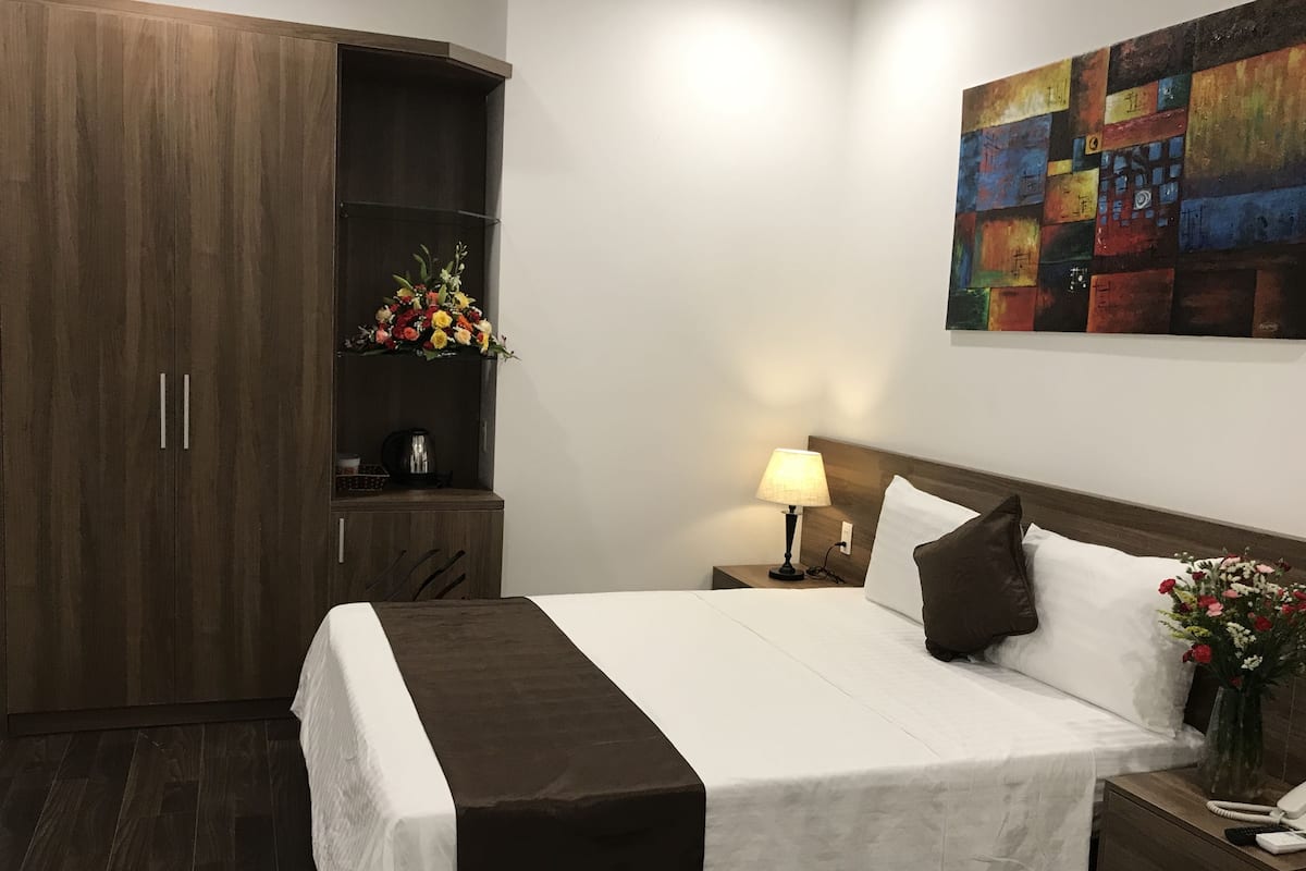 Quarto de hotel com uma cama grande, uma mesa de cabeceira com abajur, um pequeno armário com um arranjo floral e um quadro na parede.