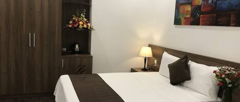 Quarto de hotel com uma cama grande, uma mesa de cabeceira com abajur, um pequeno armário com um arranjo floral e um quadro na parede.