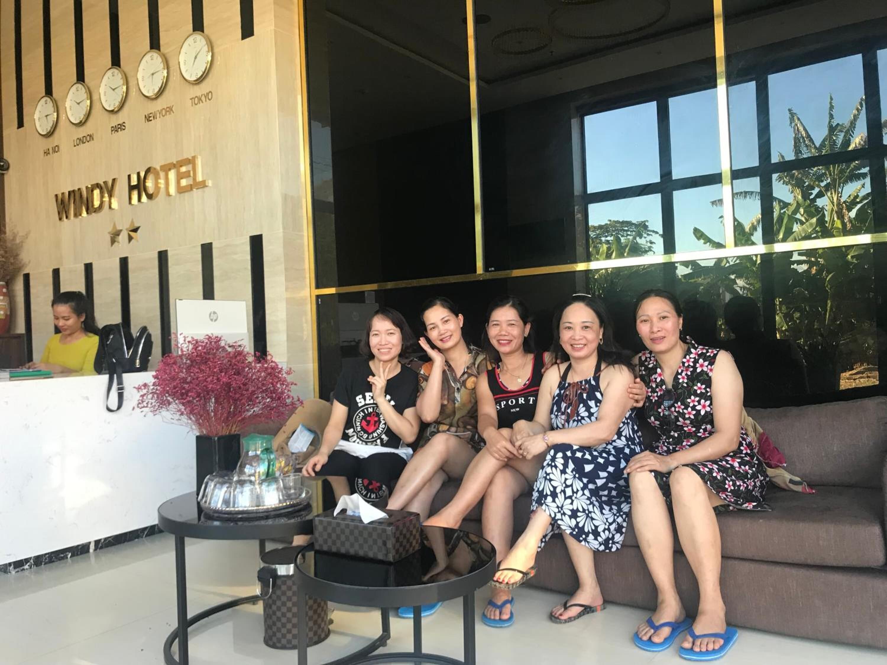 Un grupo de personas sentadas en un sofá en el vestíbulo de un hotel, con un mostrador de recepción, relojes mostrando horarios de diferentes ciudades y un gran ventanal con vistas a palmeras.