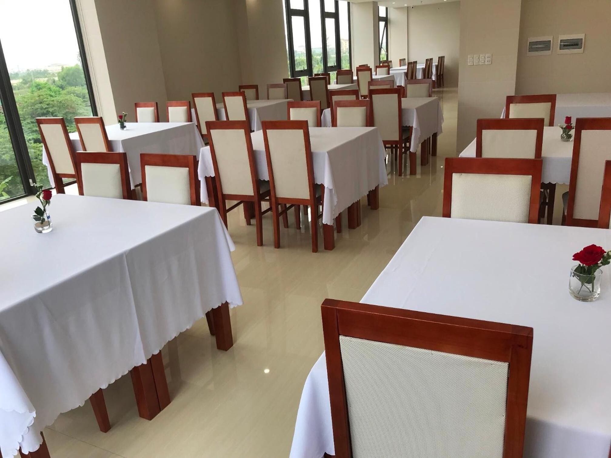 A hotel dining area with multiple tables and chairs, large windows providing a view of greenery outside.