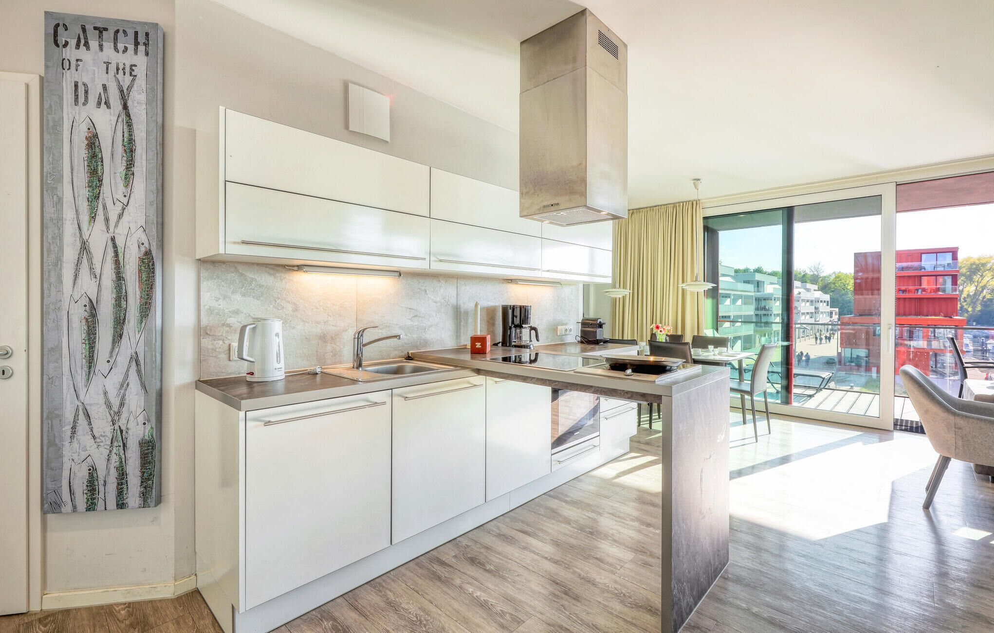 A modern kitchen with white cabinets, a stainless steel sink, and a coffee machine. There is a large window with a view of buildings outside.