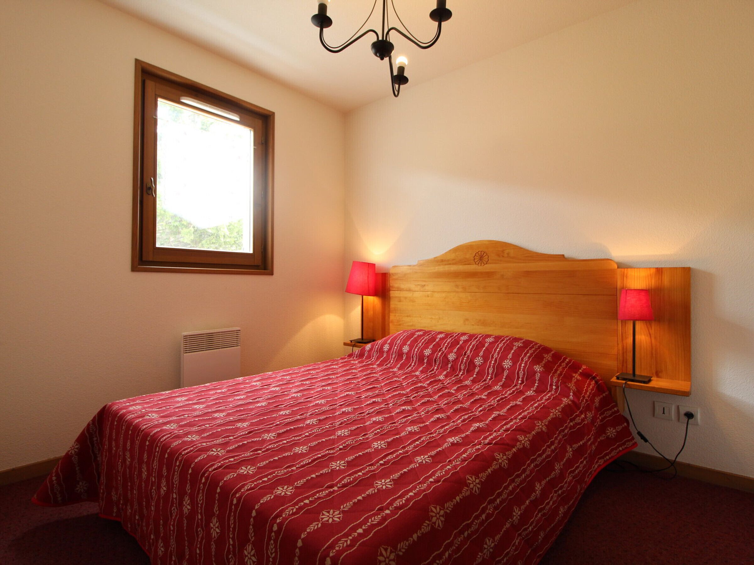 A bedroom with a wooden headboard, a red patterned bedspread, two bedside lamps, a window, and a radiator.