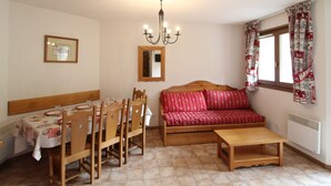 Un comedor con una mesa y sillas de madera, un sofá con cojines estampados, una lámpara de araña y una ventana con cortinas.