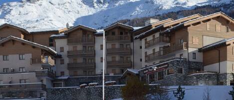 A snow-covered mountain resort with multi-story buildings, balconies, and a clear view of the mountains in the background.