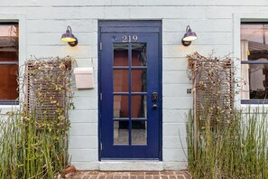 Una puerta azul con el número 219, flanqueada por dos lámparas de pared y rodeada de plantas trepadoras.