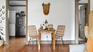 Un comedor con una mesa de madera y dos sillas de mimbre, un adorno de pared tejido, y una cocina visible a través de un vano abierto.
