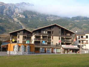 Um hotel com vários andares, varandas, uma loja de passes de esqui e montanhas ao fundo.